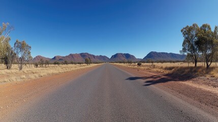 A wide-angle photo of the empty road in Australia's Outback, with red earth and sparse vegetation on both sides, a clear blue sky, and distant hills in the background.