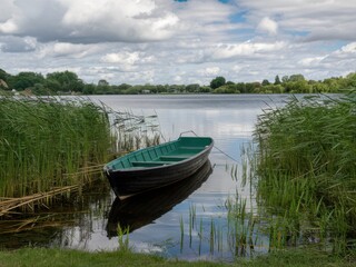Obraz premium Serene lakeside landscape, green rowboat moored in reeds, reflective calm water, lush green trees and grass, puffy white clouds, summer day, peaceful countryside scene, photorealistic, high detail, so