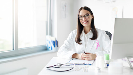 Young European doctor woman wearing pink cancer ribbon smiling at camera, working in medical office...