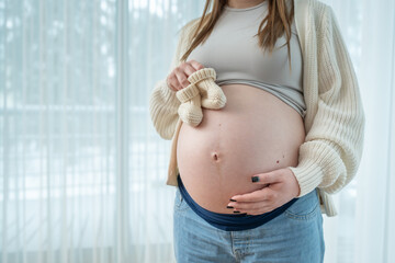 Young pregnant woman holding small knitted baby shoes on her belly near window at home in third...