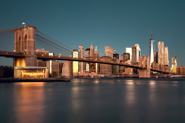 Fototapeta premium Brooklyn Bridge and Lower Manhattan skyline seen from Pebble Beach before sunrise with skyscrapers enlightened by sun and dark sky, New York City, New York, United States of America, North America.