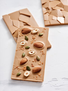 A chocolate bar features an assortment of almonds, banana chips, and pumpkin seeds, displayed attractively on a textured surface. This treat is ideal for those seeking a nutritious indulgence