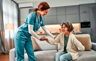 Obraz premium Geriatric doctor at home or in rehabilitation center. Nurse in blue uniform helps elderly patient to get up from sofa. Caregiver takes care of woman. Care, health care and assistance.