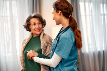 Young female doctor leaning towards smiling elderly patient, holding her hand in her palms. Doctor giving support and care. Healthcare, medicine concept. Elder care.