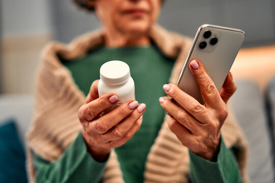 Cropped image of an elderly woman holding a bottle of pills in one hand and a phone in the other.