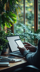 Close-up of hands holding a mobile phone with a blank white screen and a laptop on a table in the office, working online concept. Text space.