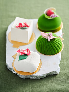 Three exquisite desserts are arranged on a marble tray, showcasing vibrant colors and delicate floral accents. Each dessert features unique shapes and textures, making them visually striking