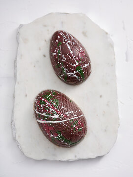 Two beautifully crafted chocolate eggs with vibrant colors and patterns are arranged on a white marble slab. These treats are ideal for holiday festivities and gatherings