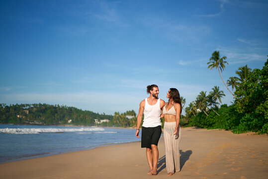 Happy couple strolls on tropical beach at sunny day. Man and woman walking barefoot by sea, enjoying vacation. Romantic walk on seaside, love, leisure time. Travelers explore coastal landscape.