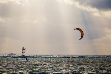 
A kitesurfer riding the sea using the power of the wind, with a ship and a sky with clouds behind him.