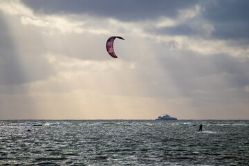 
A kitesurfer riding the sea using the power of the wind, with a ship and a sky with clouds behind him.