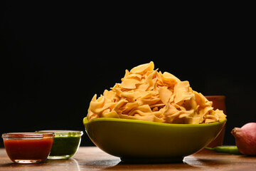 Papdi is an Indian dish that is an appetizer. When eating papdi, it is eaten with green chutney sauce along with some chili and onion. It is arranged on a dark coloured background. 