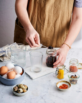A person in an apron carefully spoons ingredients from a jar while surrounded by eggs, spices, and measuring tools on a kitchen countertop