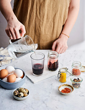 A person pours liquid from a beaker into a glass, surrounded by jars of natural dye, spices, and fresh eggs on a marble countertop, creating a festive atmosphere for egg decoration