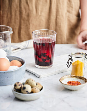 A colorful dying solution featuring dark berries sits on a marble countertop surrounded by fresh eggs, spices, and small bowls of seasonings, creating a vibrant kitchen setting