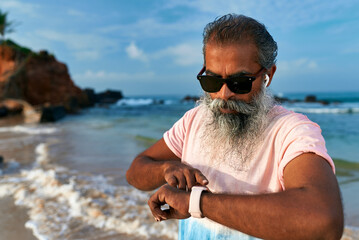 Stylish senior with gray beard at beach uses smart watch and wireless earbuds. Elderly man enjoys...
