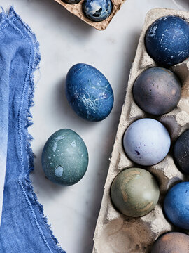 Various handcrafted easter eggs in shades of blue and green are placed in a carton on a marble surface next to a blue cloth, showcasing unique, natural designs perfect for spring celebrations