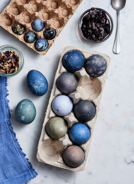 Colorful eggs in shades of blue and green are arranged in a carton alongside jars of natural dyes and dried flowers, showcasing an artistic crafting process