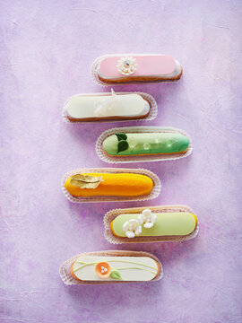 A delightful arrangement of six colorful eclairs in various flavors sits elegantly on a soft purple background. Each eclair features unique floral decorations and vivid colors