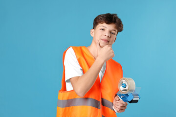Teenage boy with tape gun dispenser in safety vest working as warehouse packer on blue background