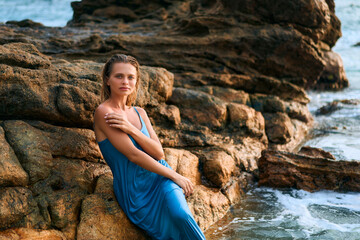 Model woman poses in wet dress by ocean rocks on exotic island. Scenic backdrop with stormy sea, vibrant landscape. Elegance in natural setting. Ideal for travel, fashion, tropical themes.