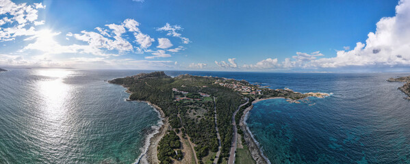 Aerial view of Capo Testa, Sardinia, showcasing rugged cliffs, lush greenery, and surrounding blue waters under a bright, sunlit sky with scattered clouds.