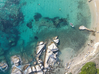 Aerial view of Zia Culumba Beach at Capo Testa, showcasing turquoise waters, sandy shoreline, scattered rocks, and a small boat anchored near the coast.