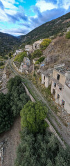 Aerial view of the abandoned village of Gairo Vecchio in Sardinia, featuring decaying stone houses overgrown with vegetation, nestled in a mountainous, green valley under a partly cloudy sky.