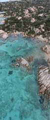 Aerial view of Caprera Island reveals crystal-clear turquoise waters surrounding rugged granite rock formations and secluded sandy coves, blending into dense Mediterranean vegetation.