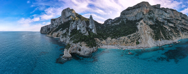 Punta Goloritzè, on the Costa di Baunei, features striking limestone cliffs rising above crystal-clear turquoise waters. The rugged coastline and serene sea blend under a vibrant sky.