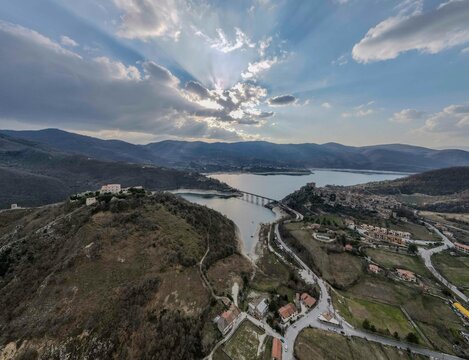 Aerial view of Lake Turano with Castel di Tora and Monte di Tora on one side and Borgo Monte Antuni and Colle di Tora on the opposite shore. The scene is framed by surrounding hills under dramatic sky
