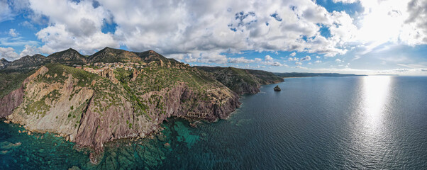 Panoramic shot of the historic mining village of Nebida in Sardinia, nestled in dramatic cliffs overlooking the vast, sparkling Mediterranean Sea under a partly cloudy sky.