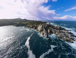 Dramatic aerial view of Capo Testa with the lighthouse, surrounded by rugged rocks and the deep blue, undulating sea under a partly cloudy sky in Sardinia.