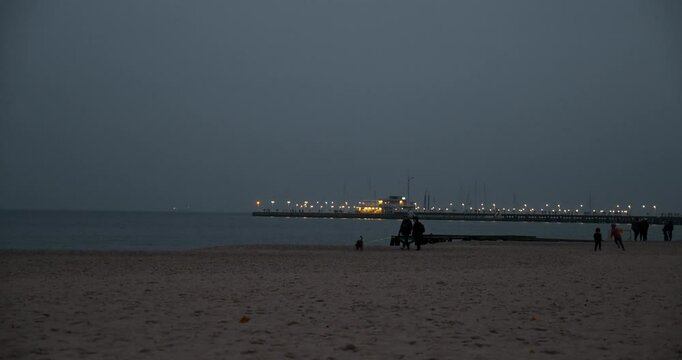 View of a beach at night with the sea, walking visitors, and a lit-up pier in the background