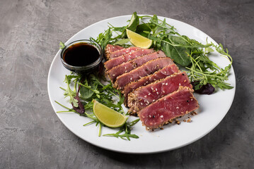 Grilled tuna steak with sesame seeds, cut into pieces with arugula and lime on a white plate on a grey background, side view.