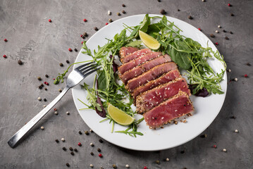 Grilled tuna steak with sesame seeds, cut into pieces with arugula and lime on a white plate on a grey background, side view.