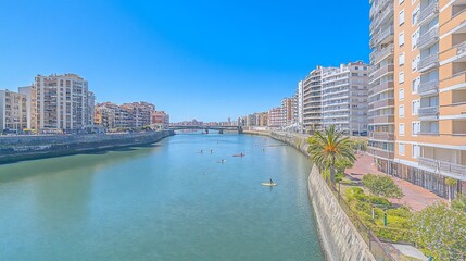 Fototapeta premium Kayakers on a calm river, flanked by modern buildings under a clear blue sky.