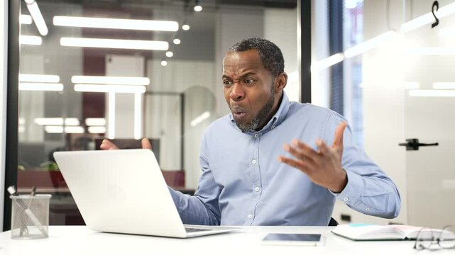 Upset worried african american businessman reads bad news on laptop while sitting at desk at workplace in business office. Frustrated worker is sad while reviewing negative information on a computer