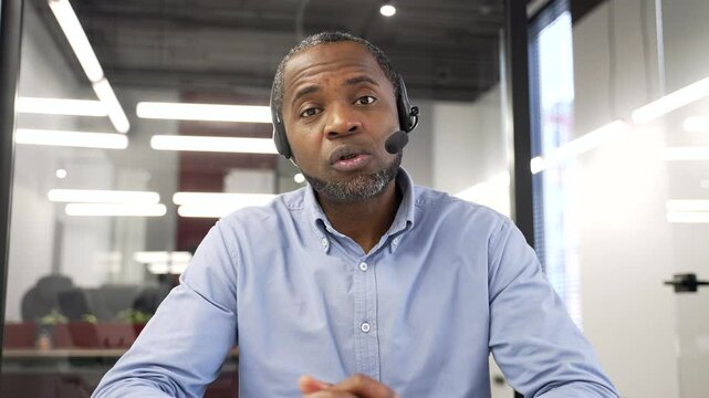 Confident african american businessman in wireless headset talking on a video call looking at camera sitting in office. Entrepreneur has a business meeting. Coach speaks remotely at online training
