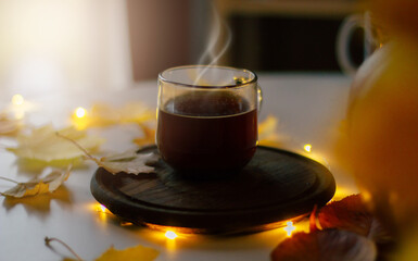 A steaming cup of coffee sits on a wooden coaster surrounded by autumn leaves and soft glowing lights, creating a cozy fall atmosphere. Perfect for seasonal lifestyle and comfort-themed content.