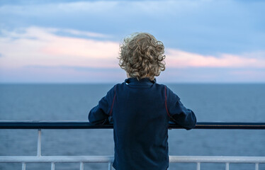A young child with curly hair stands on a ferry, resting his arms on the railing, gazing at the distant horizon. The sky displays soft hues as the sun sets over the water