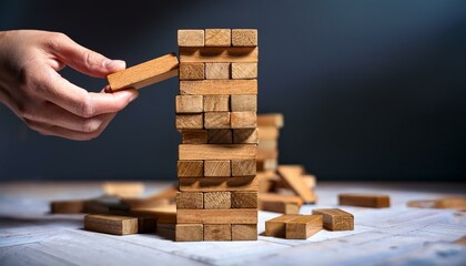 A hand lifts the last block as a precariously stacked wooden block tower is in danger of collapsing.