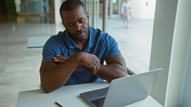 Young man in office looking exhausted with hand on head and laptop on table, indoor setting implying workplace stress