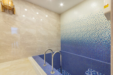 A small swimming pool decorated with mosaic tiles in blue tones. On the wall is a wooden tub for...