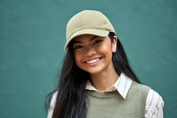 Smiling pretty gen z Asian teen girl fashion model looking at camera posing at color wall. Close up headshot portrait of happy stylish cool female mode with beautiful wide smile wearing cap.
