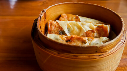 Three spring rolls or lumpia in a bamboo steamer basket, placed on a wooden table