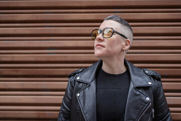 Short-haired woman wearing sunglasses and leather jacket posing near brown background