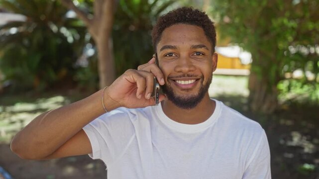 Young man talking on phone in urban park, showcasing a smiling adult black male wearing a white shirt outdoors with trees in the background