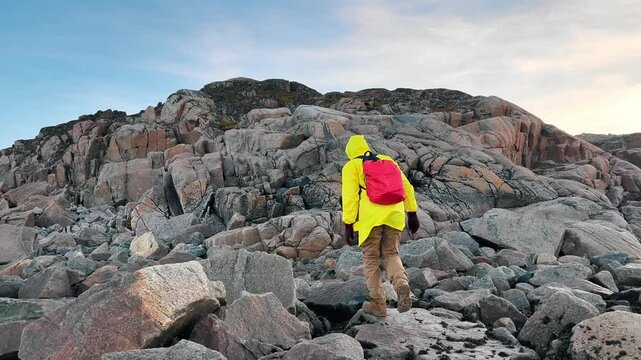 Traveler, clad in vibrant yellow raincoat, and equipped with backpack, begins ascent of hill, which is covered in diverse rocks and boulders.