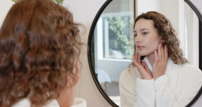 Woman in bathrobe examining skin in mirror, practicing self-care routine